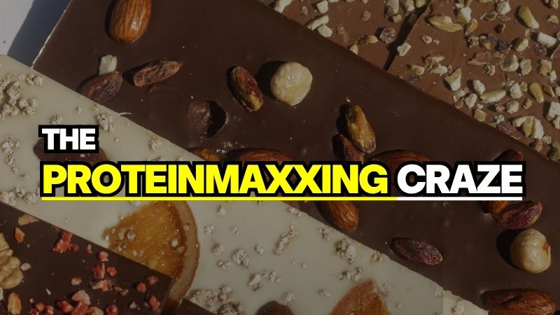 A dark chocolate bar topped with various nuts, displaying the text "THE PROTEINMAXXING CRAZE" to represent the oroteinmaxxing trend in modern food culture.
